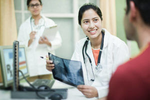 Indoor over shoulder view shot at clinic of a happy, Asian, female doctor holding an x-ray image of cervical vertebrae and talking to the male patient while junior doctor taking notes on a clipboard in the background. Three people, waist up, horizontal composition with selective focus.
