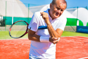 Senior man who his standing during tennis match on sunny day is experiencing elbow pain caused by tennis elbow.