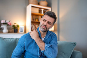 Man sitting on the sofa and holding painful shoulder with hand while suffering from rotator cuff injury.